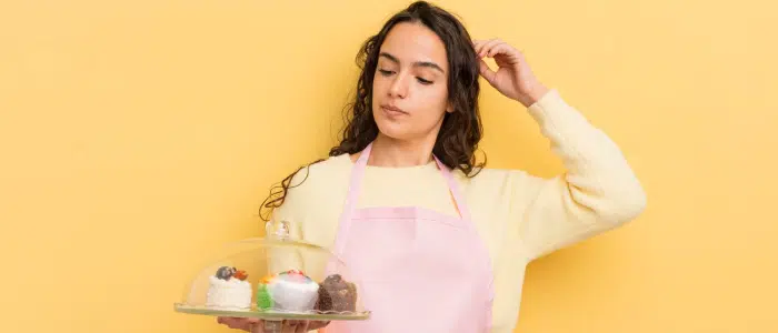 a woman in a pink apron holding a tray of cakes and looking confused, showing a common cake baking mistake moment
