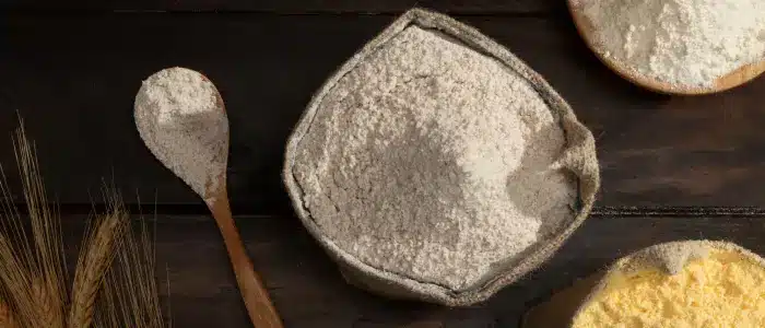 Top view of whole wheat flour in a rustic cloth bowl with a wooden spoon beside it, surrounded by various natural flours emphasizing healthy baking choices made from nutrient-rich, unrefined ingredients
