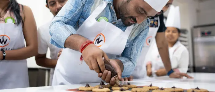 Man in a chef hat and apron carefully piping chocolate onto cookies during a baking class, showcasing precision and skill in piping techniques as part of creative bakery artistry