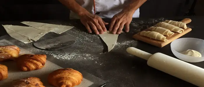 Chef folding croissant dough on a floured counter with baked pastries, rolling pin, and dough in view, highlighting the craftsmanship and essential role of baking and pastry in culinary world