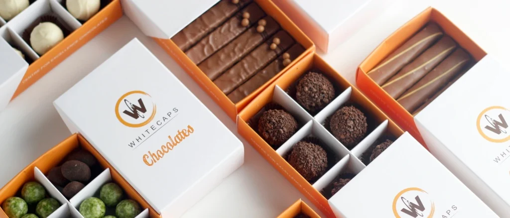 Assorted premium chocolates in elegant Whitecaps branded boxes, featuring truffles, chocolate bars, and coated nuts arranged neatly for display